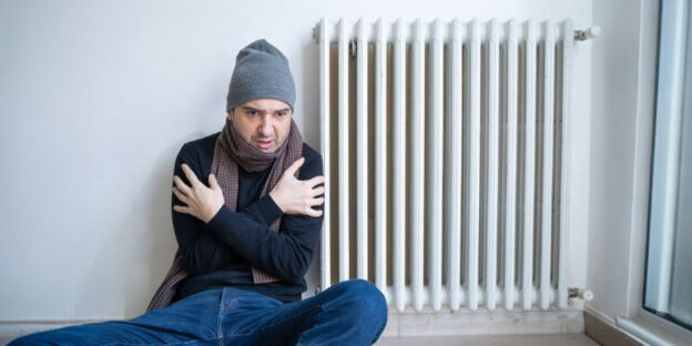 A man sitting on the floor near a radiator, trying to keep warm.